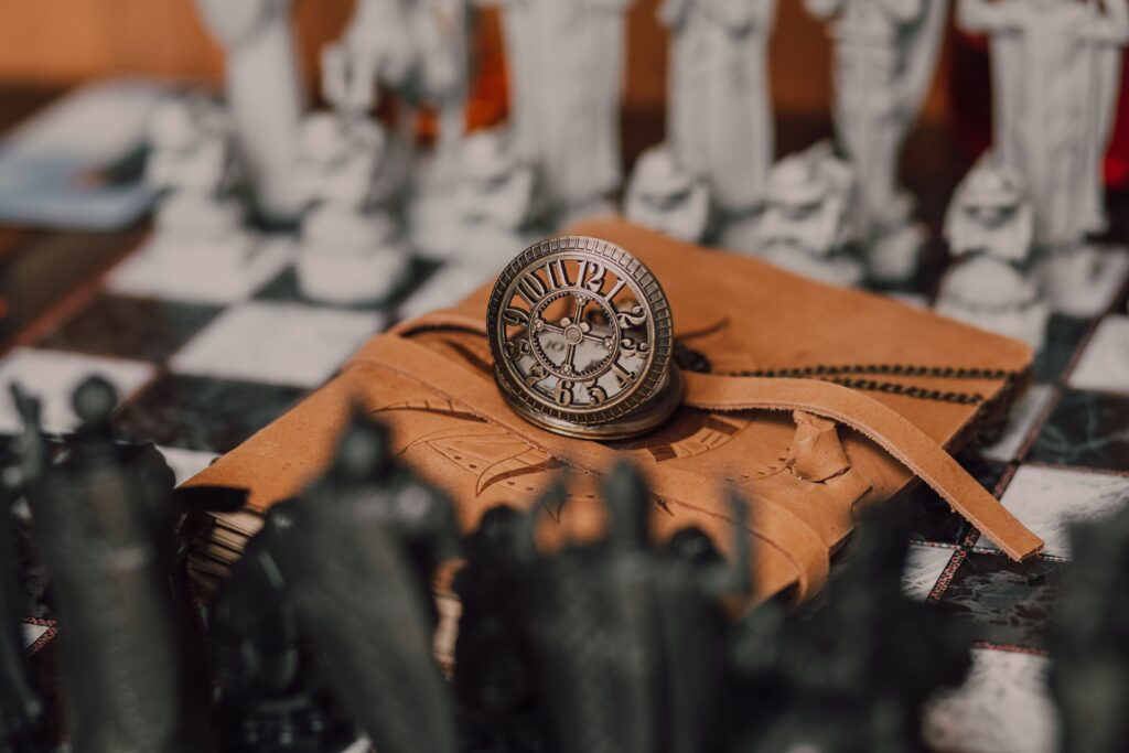 A steampunk pocket watch sits on top of a brown leather notebook, in the centre of a chess board with intricate chess pieces.